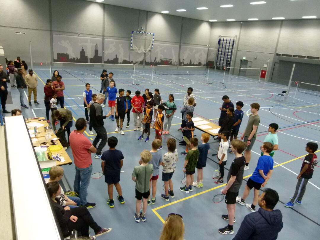 Spelers en ouders staan in de hal te luisteren naar Wims uitleg. De foto is gemaakt van bovenaf.